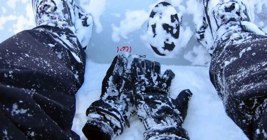 a snowboarder sitting with boots attached to the snowboard with snowboard gloves laying on snow