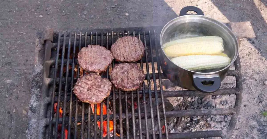 burgers and a pot with corn cooking over a campfire depicts the best campfire grill grates