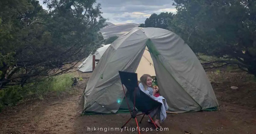 a girl sitting in a camp chair outside of a camping tent at a developed campground